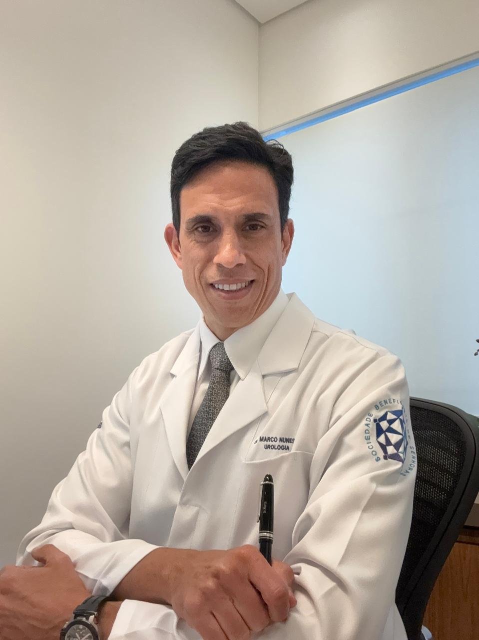 urologista-particular Urologista particular Dr. Marco Nunes sorrindo em seu consultório, usando jaleco branco com identificação e gravata cinza, transmitindo confiança e acolhimento aos pacientes.