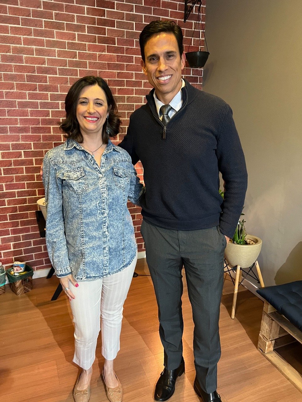 Dr. Marco Nunes e Andréa Moreira posam sorridentes após o episódio especial do podcast Novembro Azul Senhores da Barba, em ambiente interno com parede de tijolos e iluminação aconchegante.