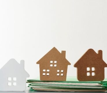 Miniaturas de casas sobre uma pilha de documentos, representando decisões de moradia e qualidade de vida