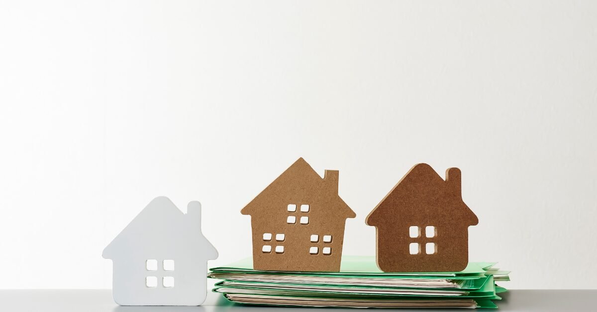 Miniaturas de casas sobre uma pilha de documentos, representando decisões de moradia e qualidade de vida