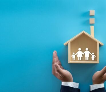 Mãos protegendo uma casa de madeira com símbolo de família, representando segurança, saúde e bem-estar no lar