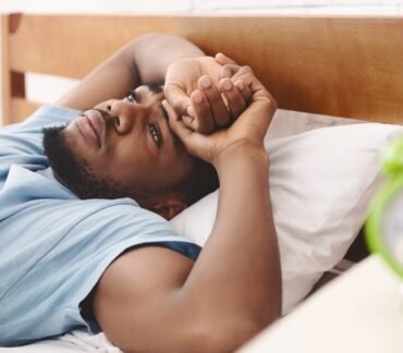 Homem deitado na cama acordado pensativo com relógio verde desfocado representando acordar à noite para urinar