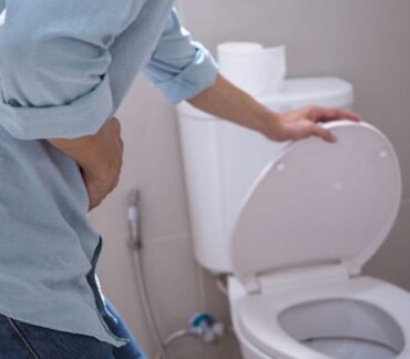 Homem no banheiro, curvado e segurando a região do abdômen, próximo ao vaso sanitário com a tampa levantada.