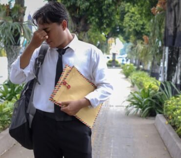 Homem de camisa social e gravata, com expressão de cansaço, segurando o rosto na calçada.