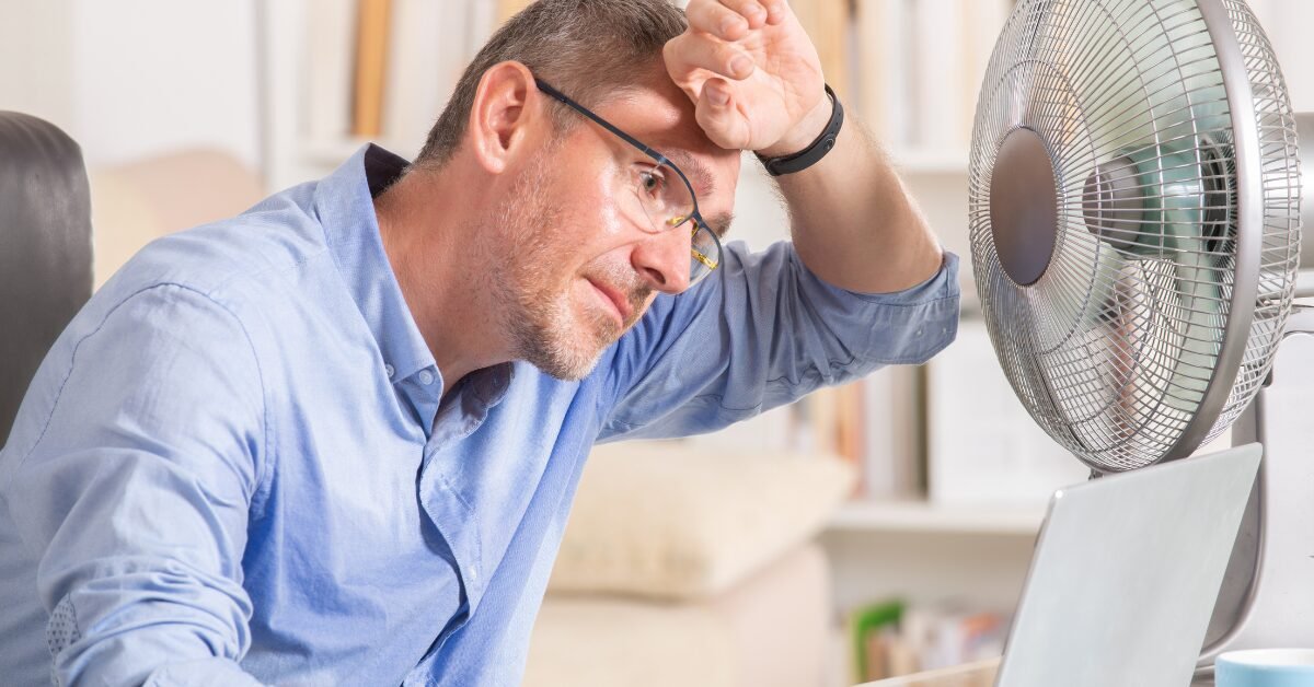 Homem de óculos suando, mão na testa, sentado em frente a um laptop com ventilador ao lado. Title: Homem suando em home office quente.